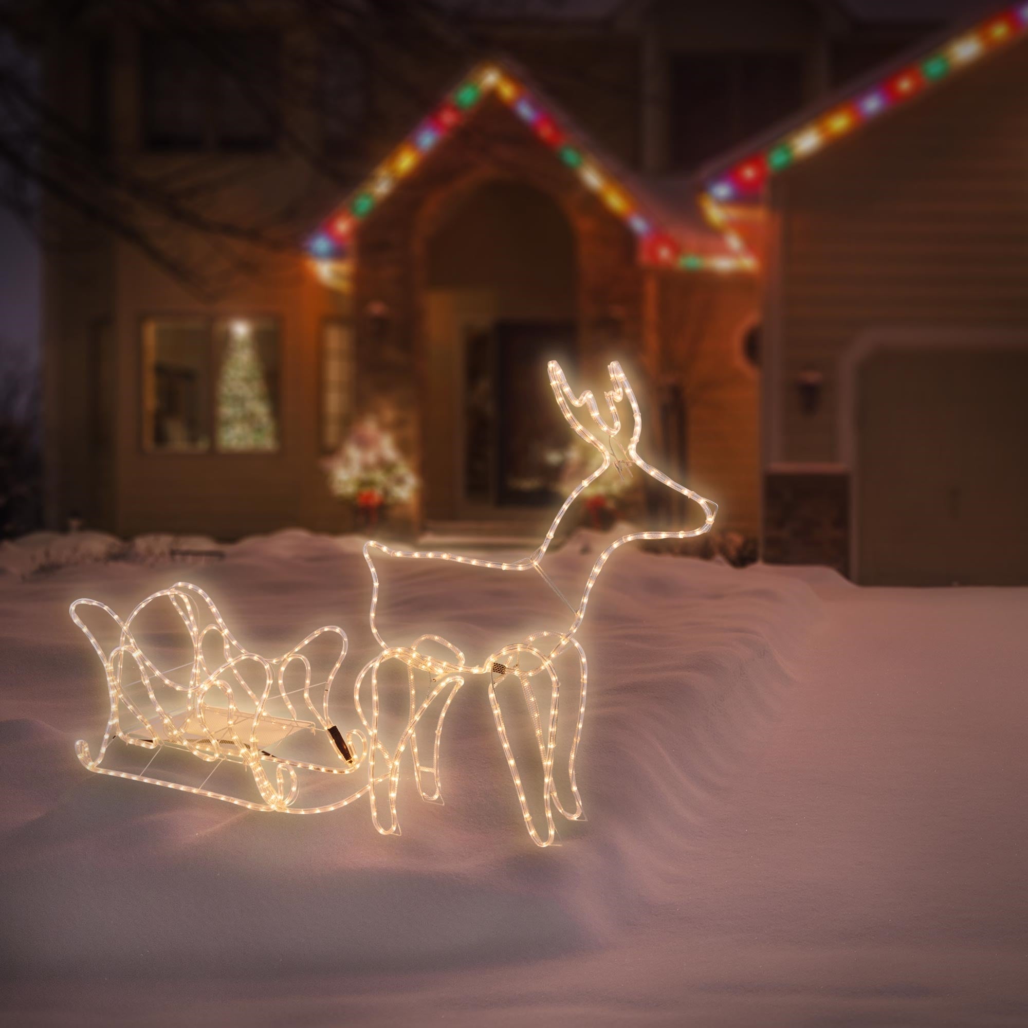Renne illuminé et traîneau dans la neige devant une maison décorée pour les fêtes.