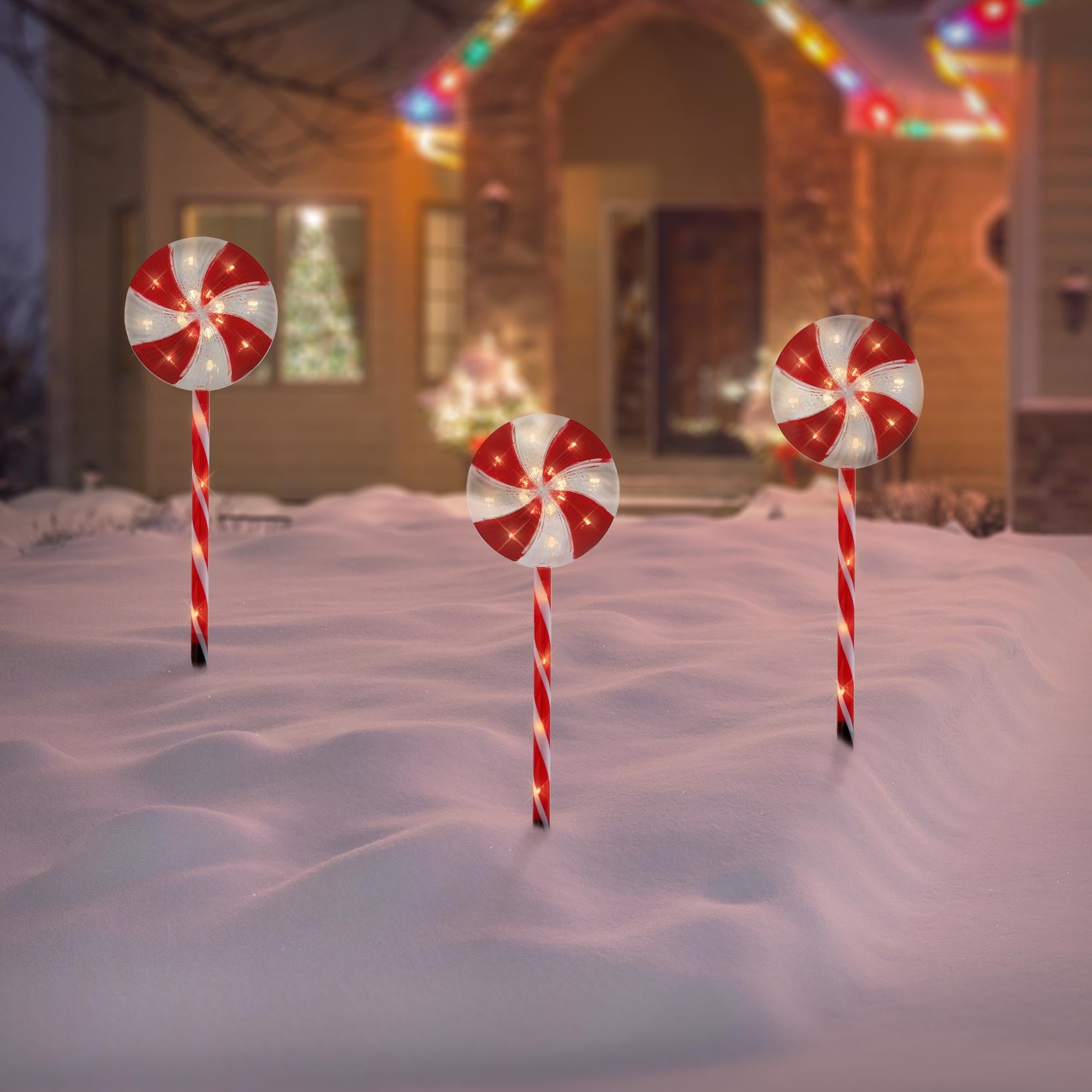 Drei große, leuchtende Lutscher stehen im Schnee vor einem festlich geschmückten Haus.