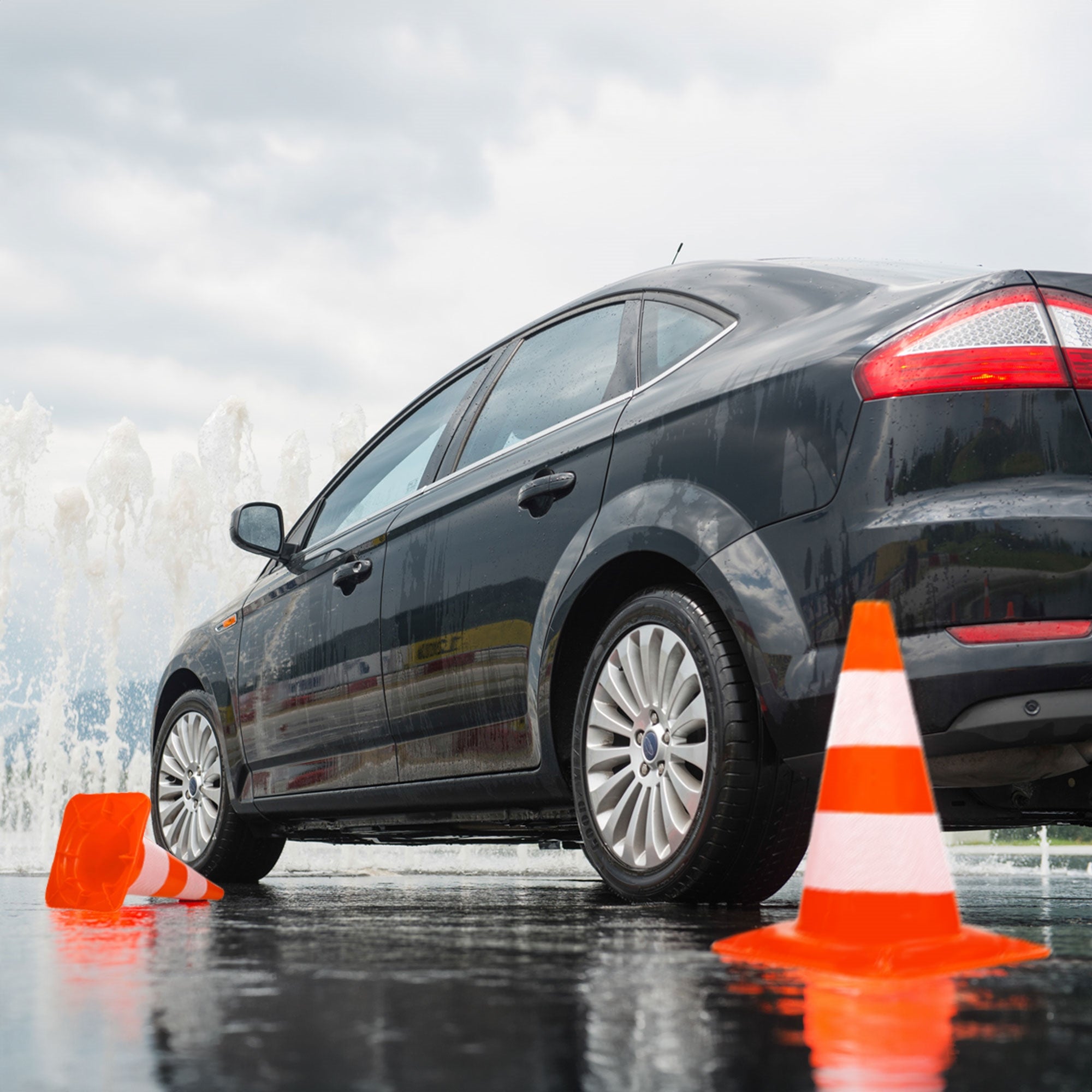 Voiture noire à côté de cônes orange sur une route mouillée avec des éclaboussures en arrière-plan.