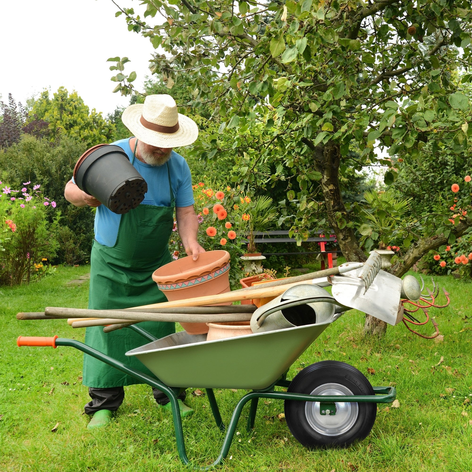 Uomo in giardino con carriola, vasi e attrezzi, circondato da fiori.