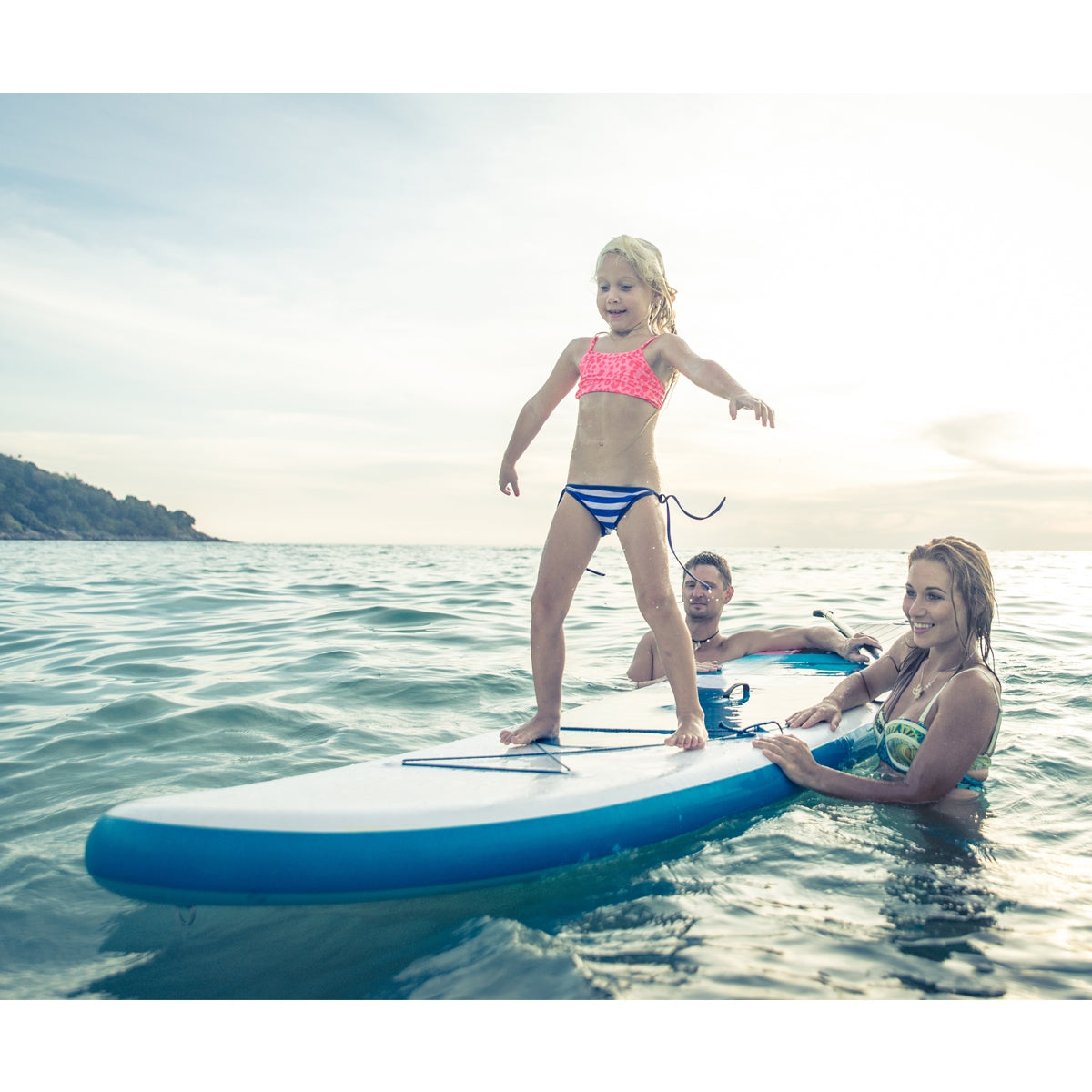 Dziecko stoi na desce surfingowej w wodzie, otoczone przez dwóch dorosłych.
