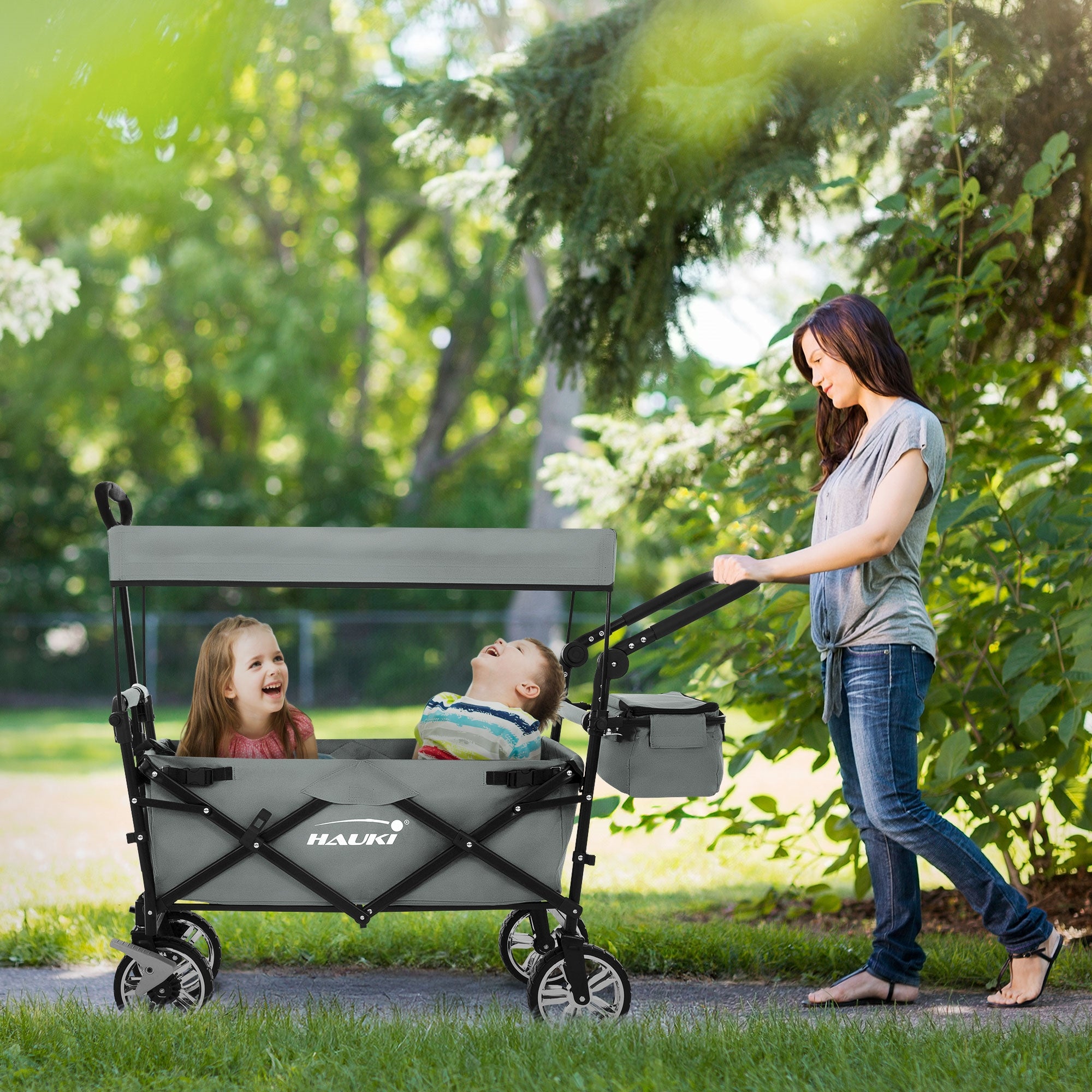 Une femme pousse une poussette grise avec deux enfants dans le parc.
