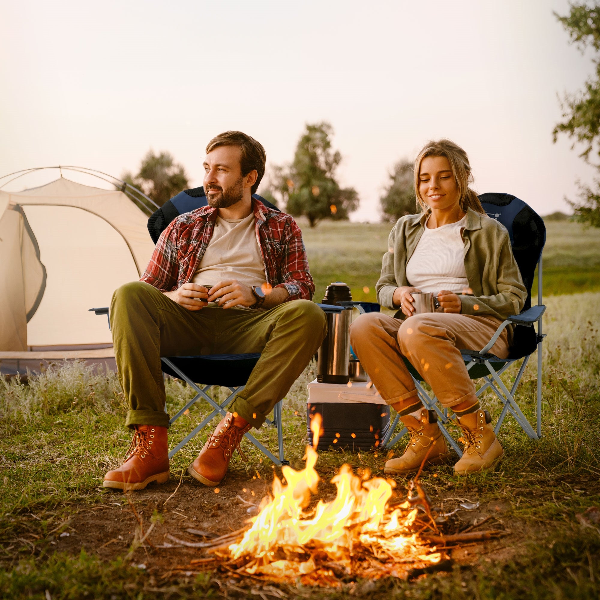 Un couple assis autour d'un feu de camp en plein air, avec une tente en arrière-plan.