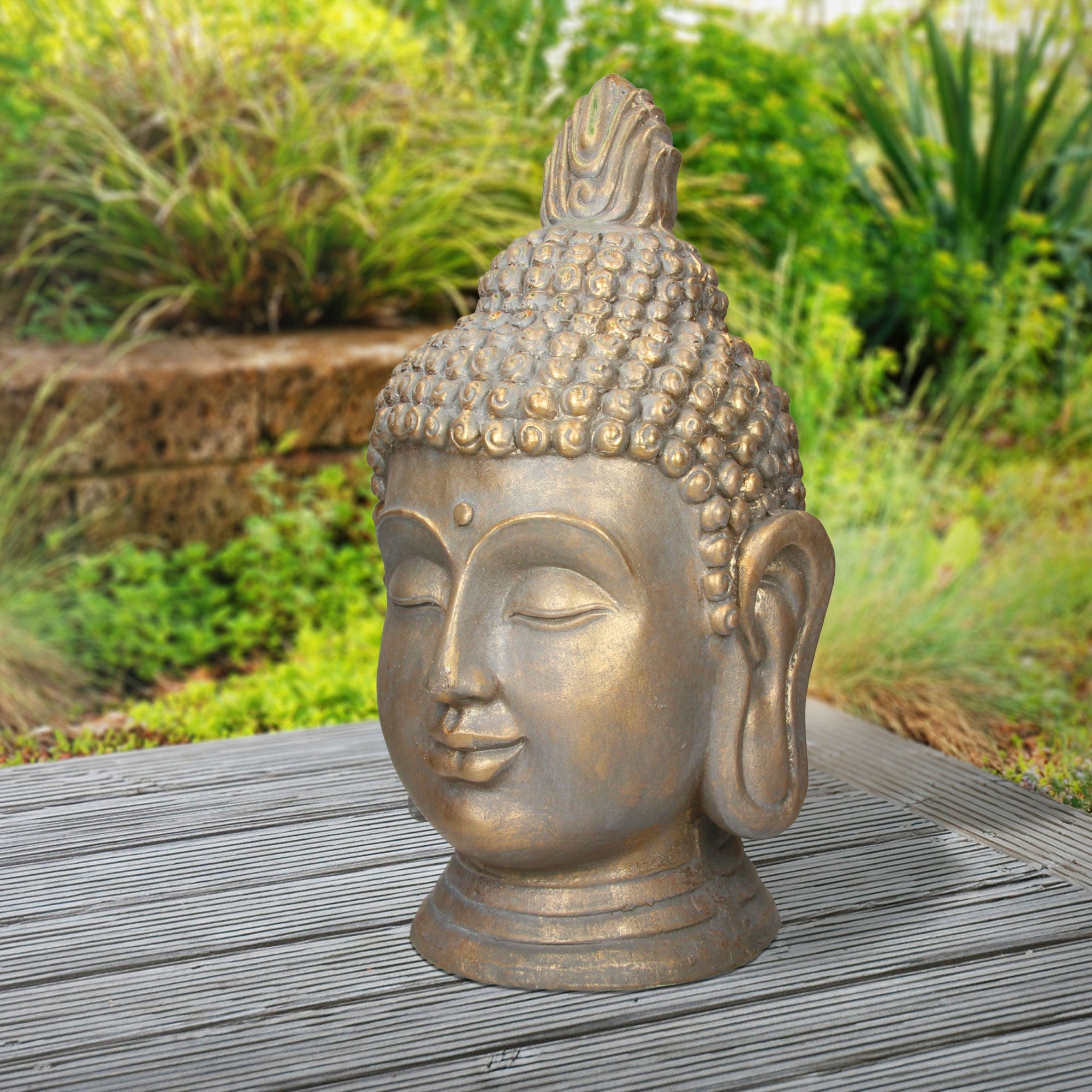 Pronssinen Buddha-pää ulkona pöydällä, ympärillä kasveja.