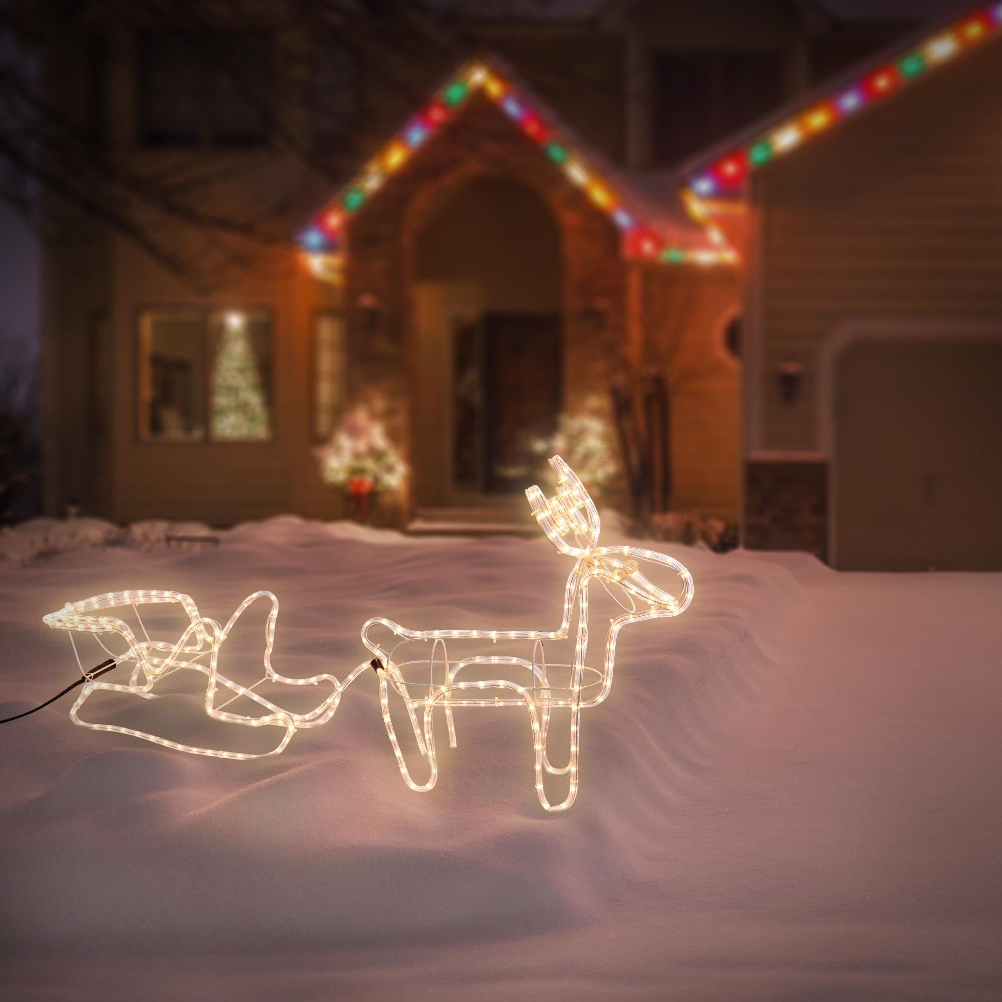 Beleuchtetes Rentier mit Schlitten im Schnee vor einem Haus.