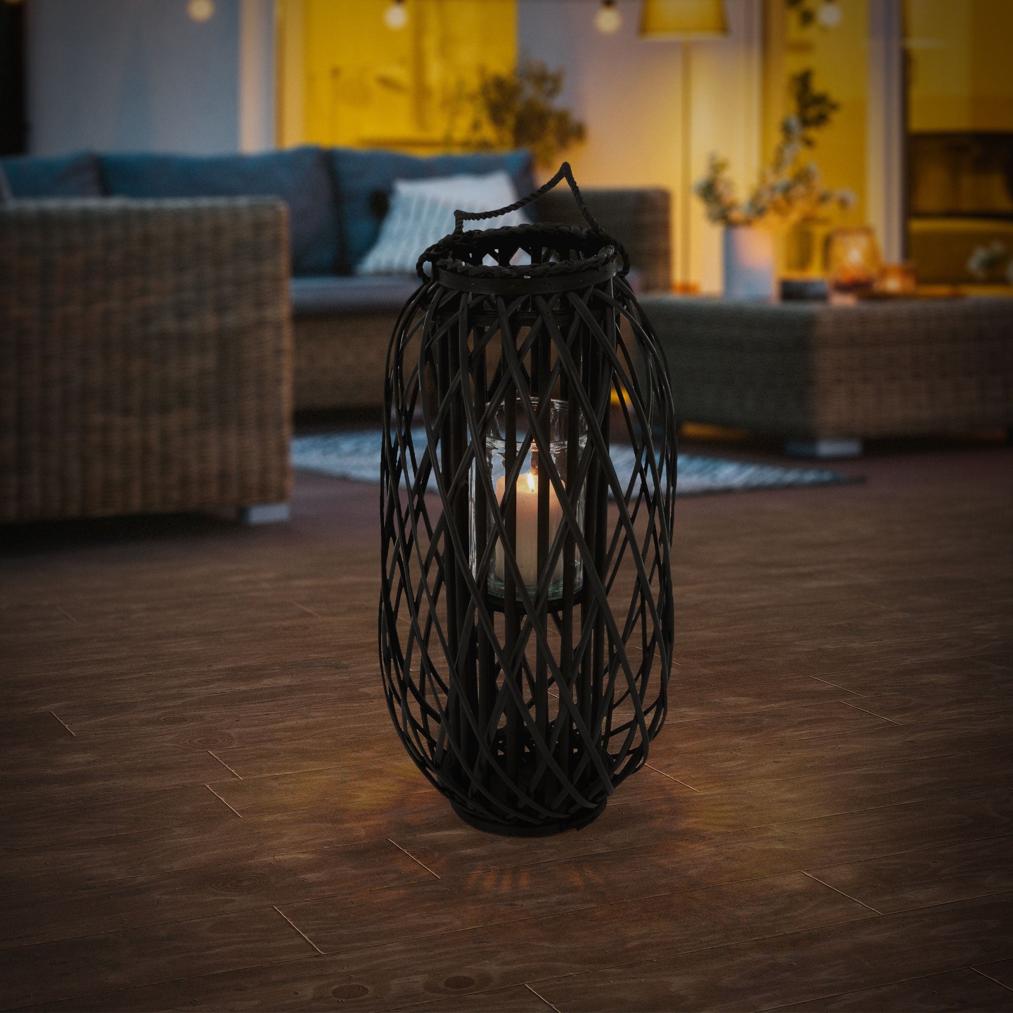 Lanterne noire en roseau avec une bougie sur un sol en bois dans une pièce confortable.