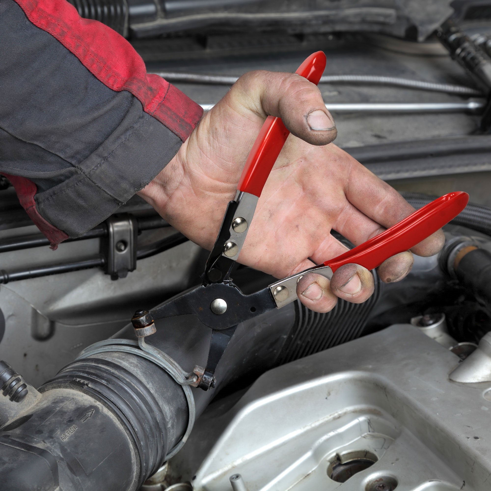 Mano con pinza sopra un vano motore, abbigliamento da lavoro rosso e nero.