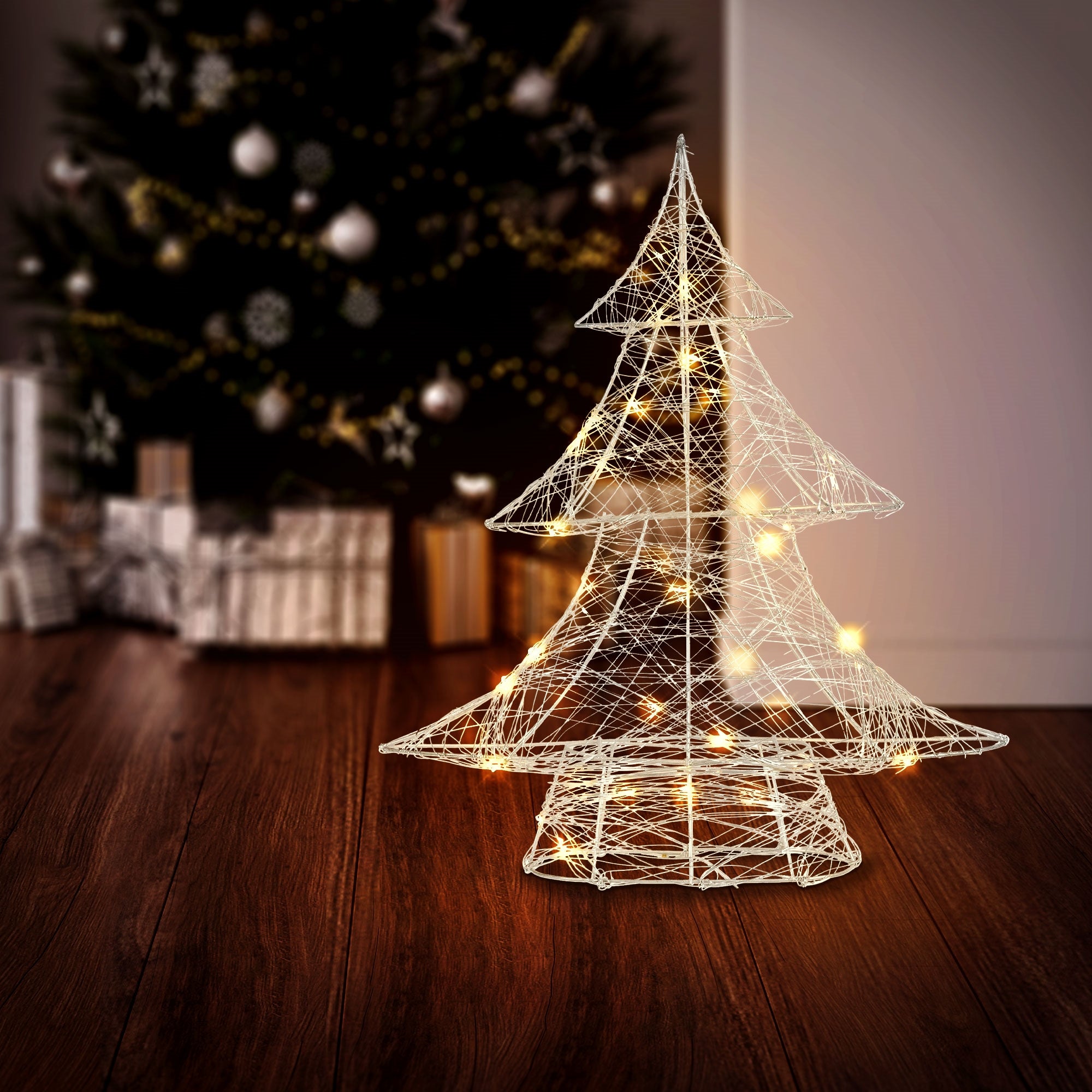 Drahtbaum mit Lichtern vor einem Weihnachtsbaum und Geschenken.