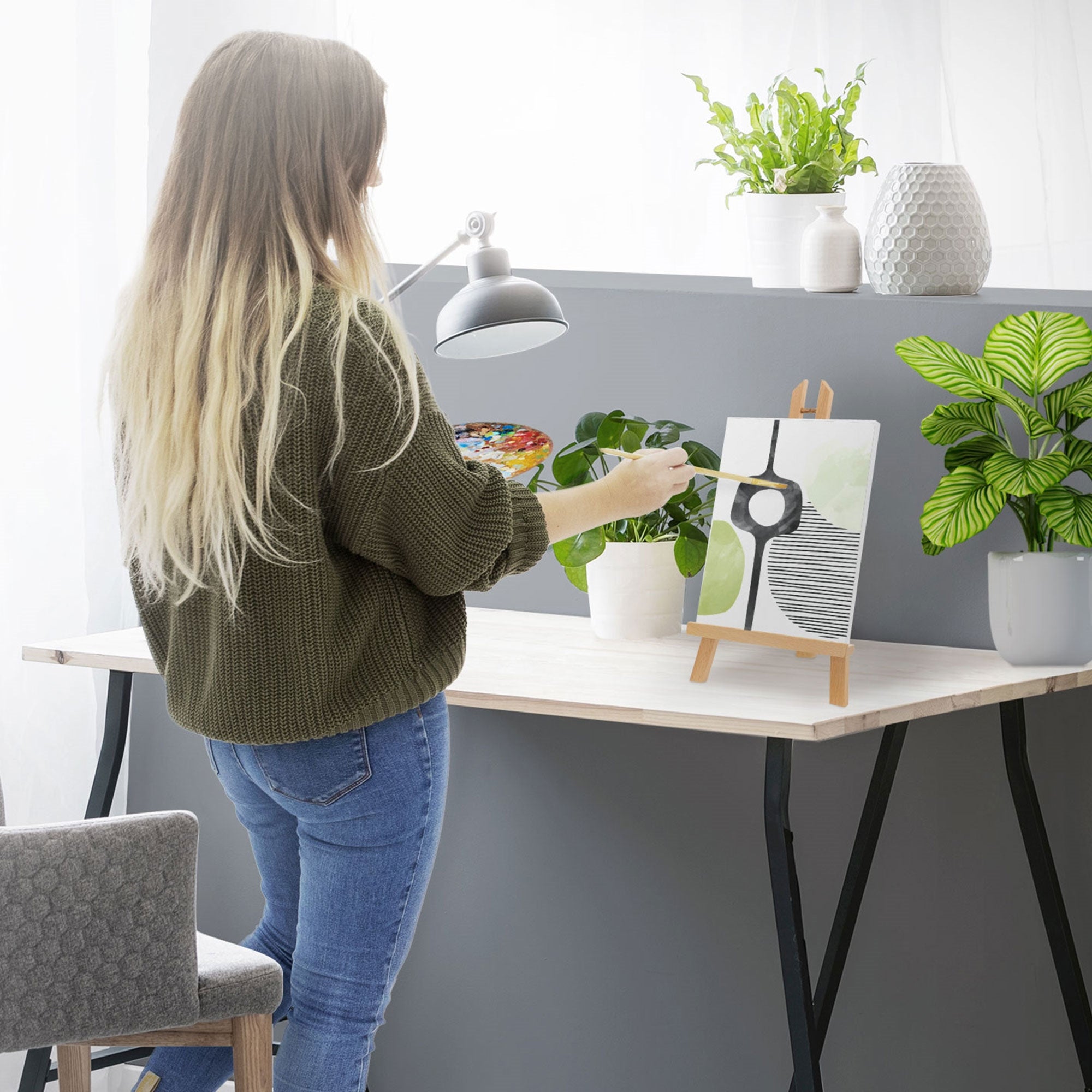 Vrouw schildert aan een tafel, kunstwerk op een standaard, planten op de achtergrond.