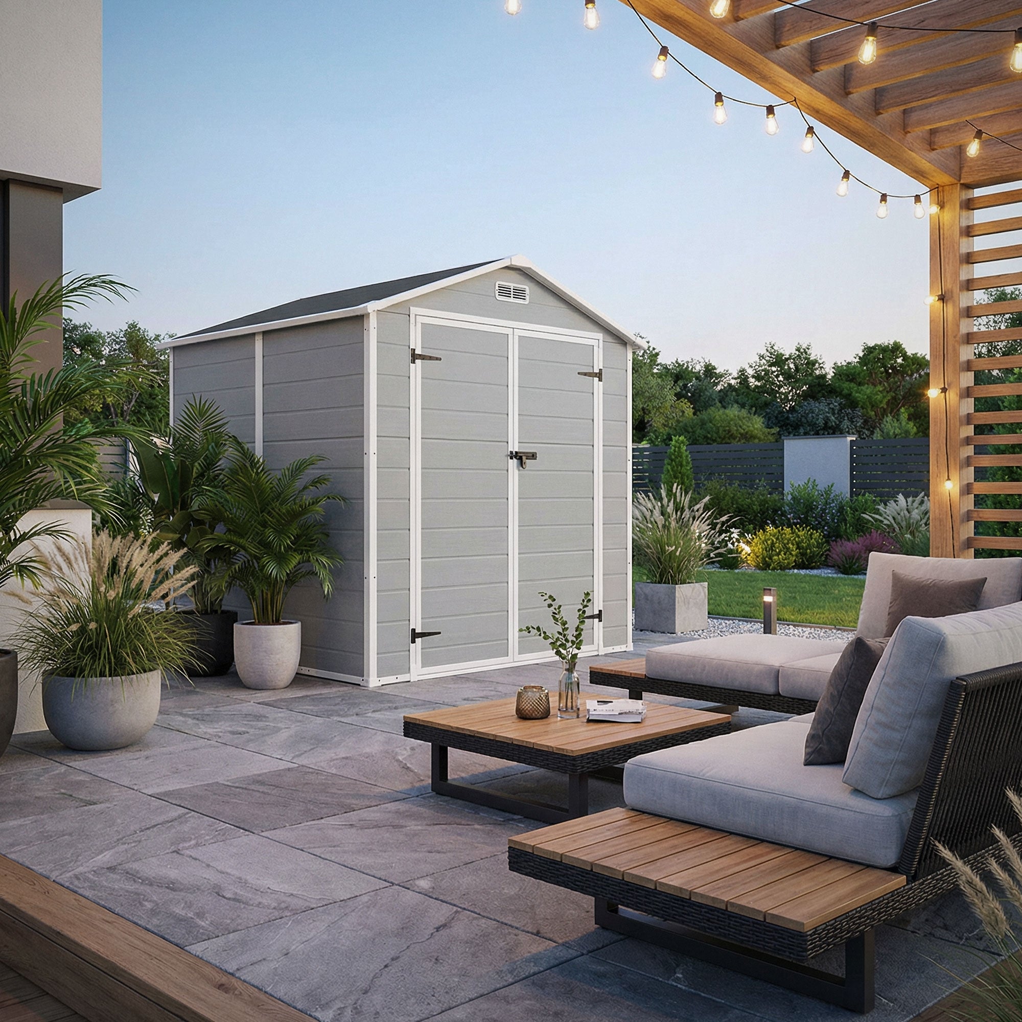 Helles Gartenhaus auf einer Terrasse, umgeben von Pflanzen und Möbeln, mit Abendbeleuchtung.