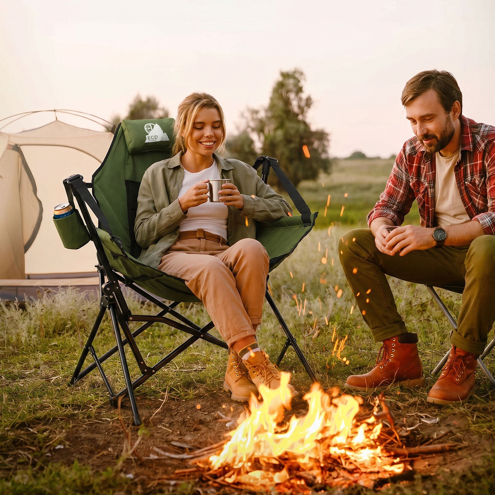 Zwei Personen sitzen am Lagerfeuer, eine hält eine Tasse, Zelt im Hintergrund.