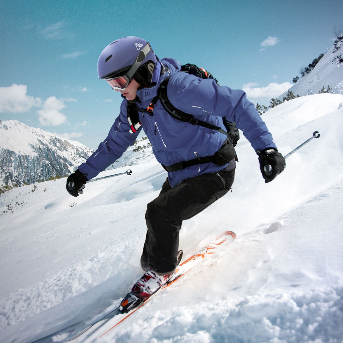Skifahrer in blauer Jacke fährt auf schneebedecktem Hang mit Bergen im Hintergrund.