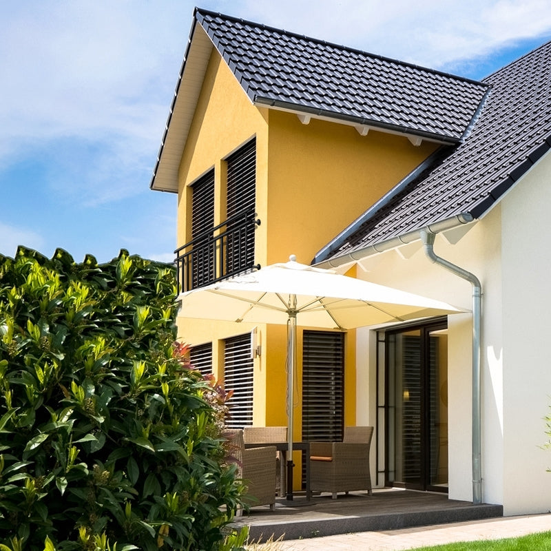 House with yellow facade, terrace, and sunshade, surrounded by plants.