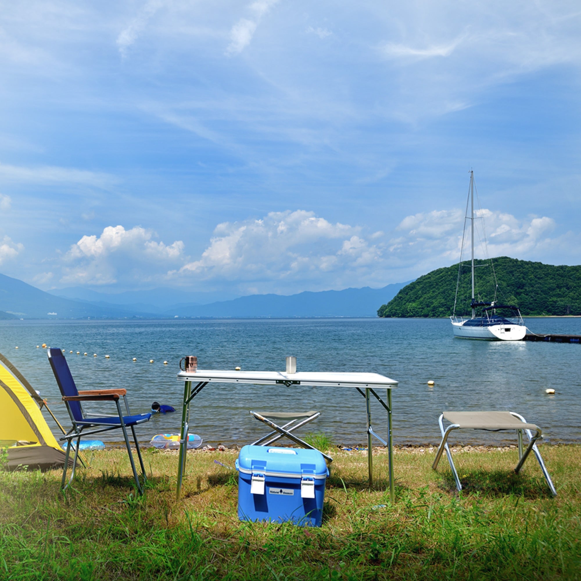 Vue sur un lac avec un bateau, des tables et des chaises sur la rive sous un ciel bleu.