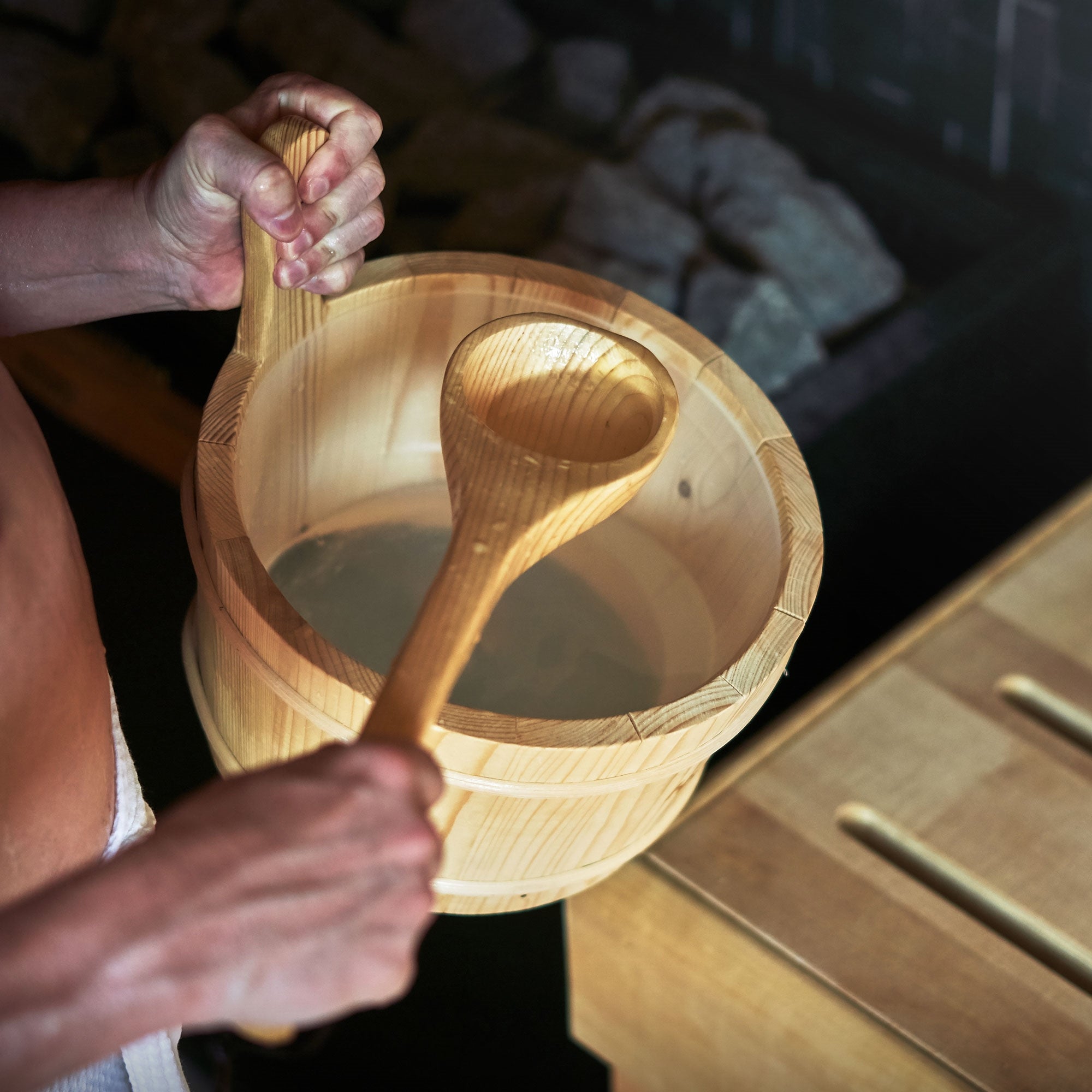 Hände halten einen Holzbehälter mit Wasser und einem Holzlöffel.