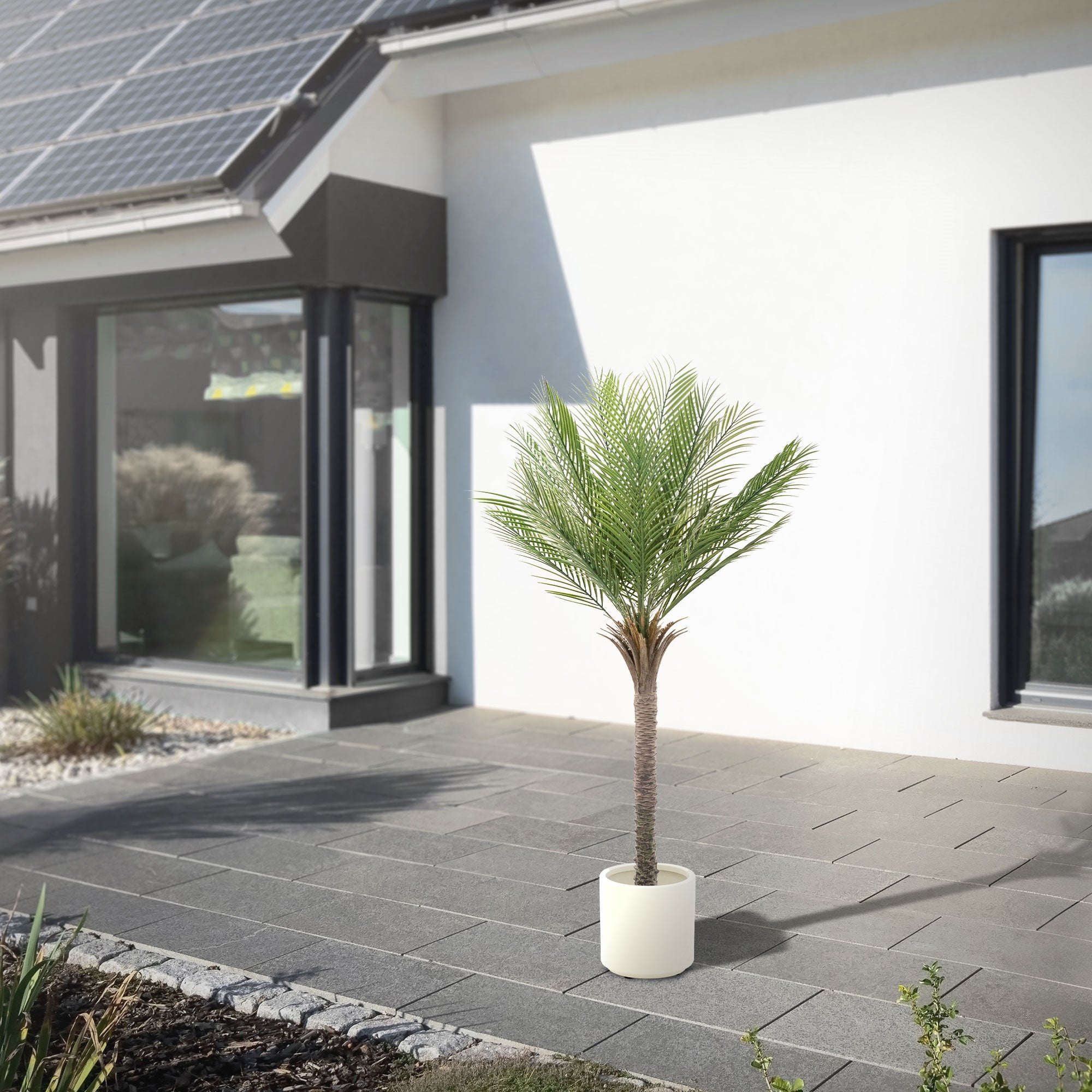 A tall artificial tree in a white pot in front of a modern house with solar panels.