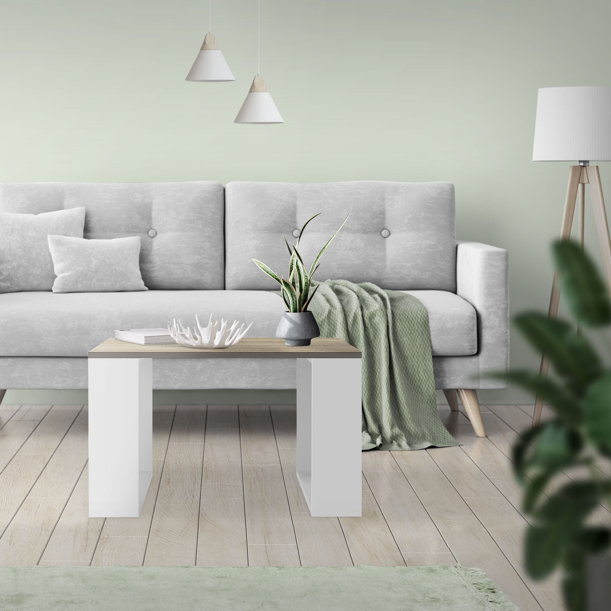 Een woonkamer met een grijze bank, tafel en planten op een lichte vloer.