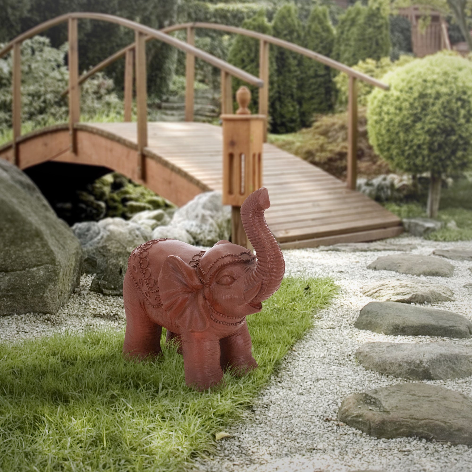 Roter Elefant aus Kunststoff im Garten vor einer Holzbrücke.