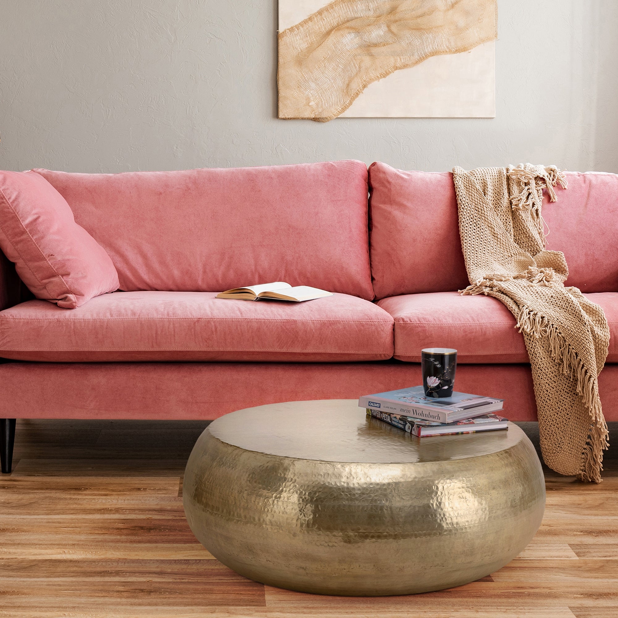 Rosa Sofa mit Kissen und einer Decke, runder Tisch mit Büchern und einem Glas.